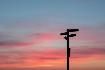 A signpost with 4 signs silhouetted against a pink and orange sunset